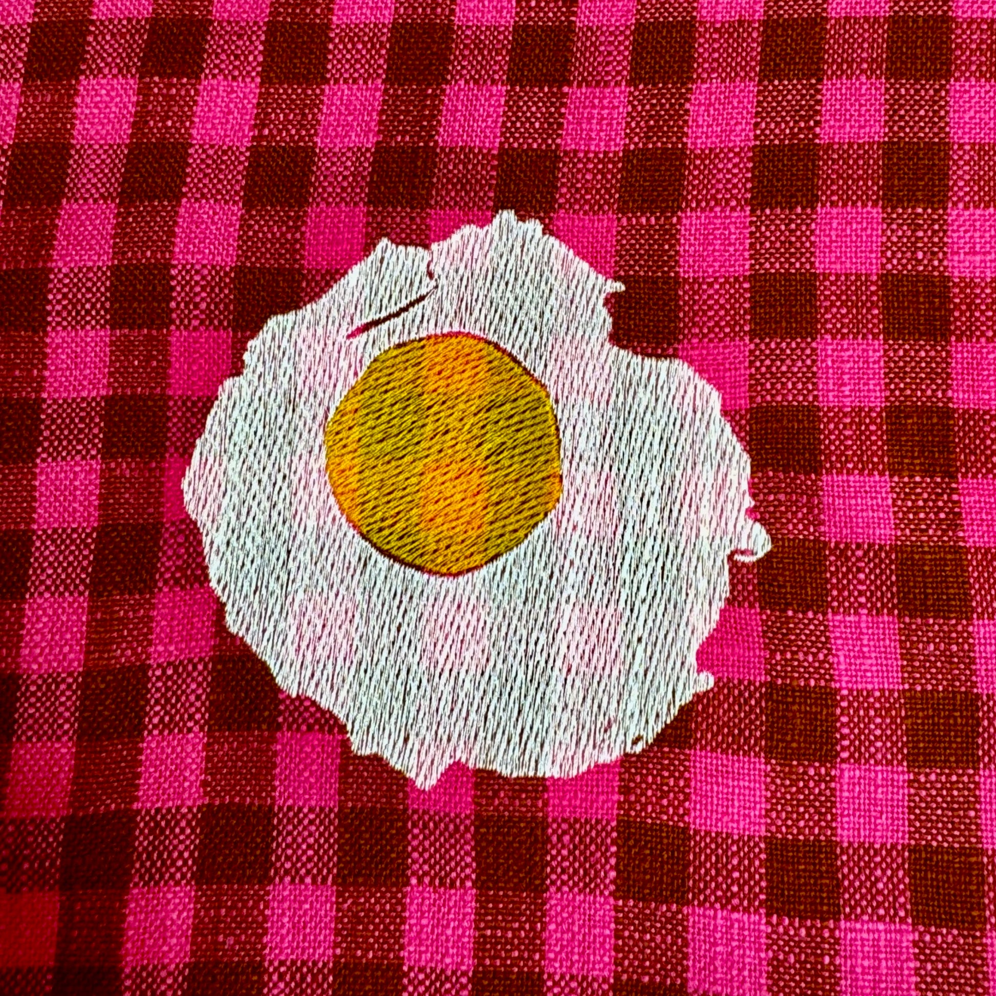 Close-up of hand-drawn sunny-side-up egg embroidery in yellow and white thread on fuchsia gingham cotton tea towel