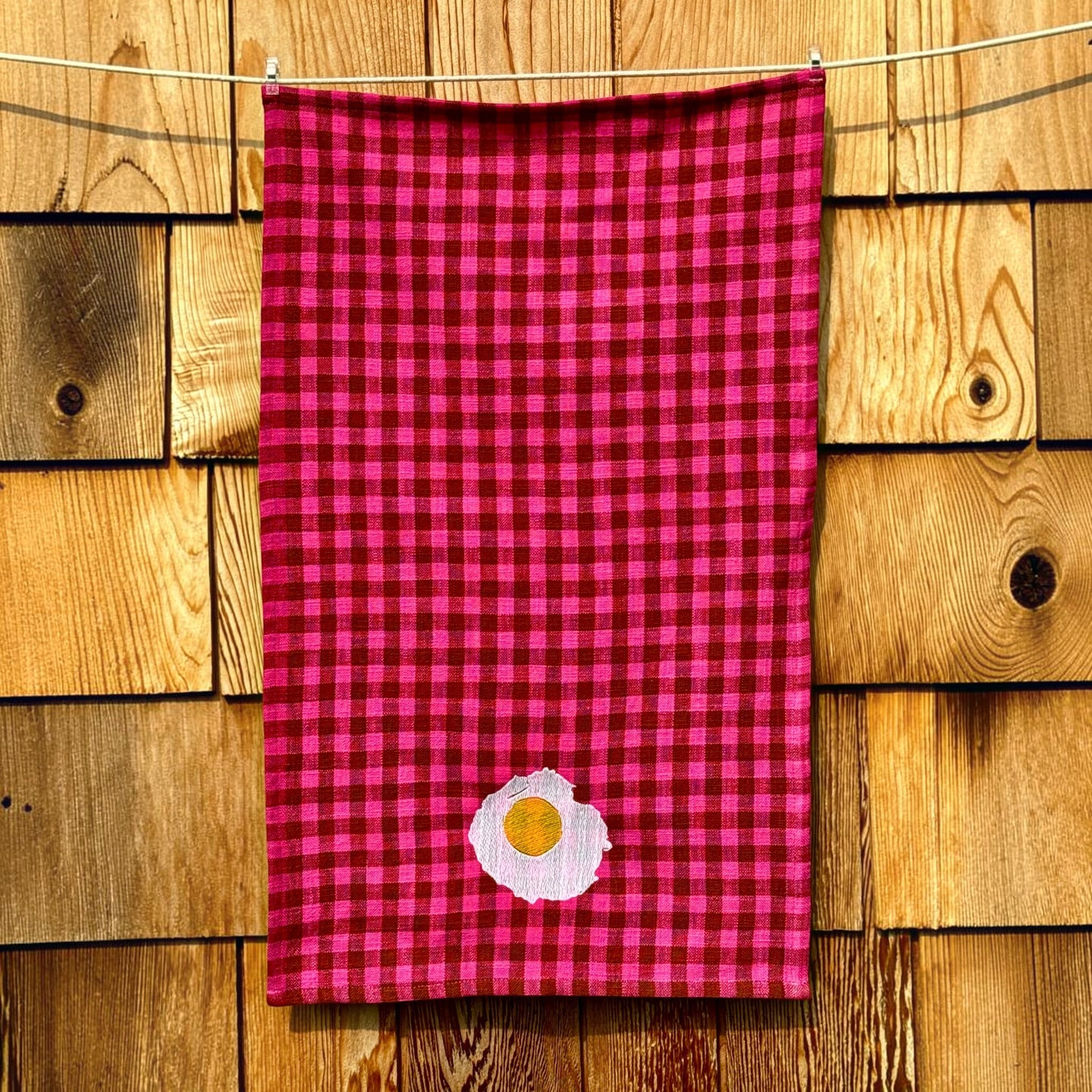 Full-length view of fuchsia and cocoa gingham tea towel with embroidered sunny-side-up egg near the hem