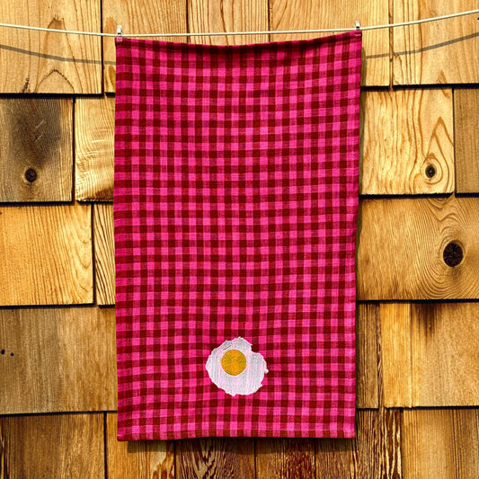 Full-length view of fuchsia and cocoa gingham tea towel with embroidered sunny-side-up egg near the hem