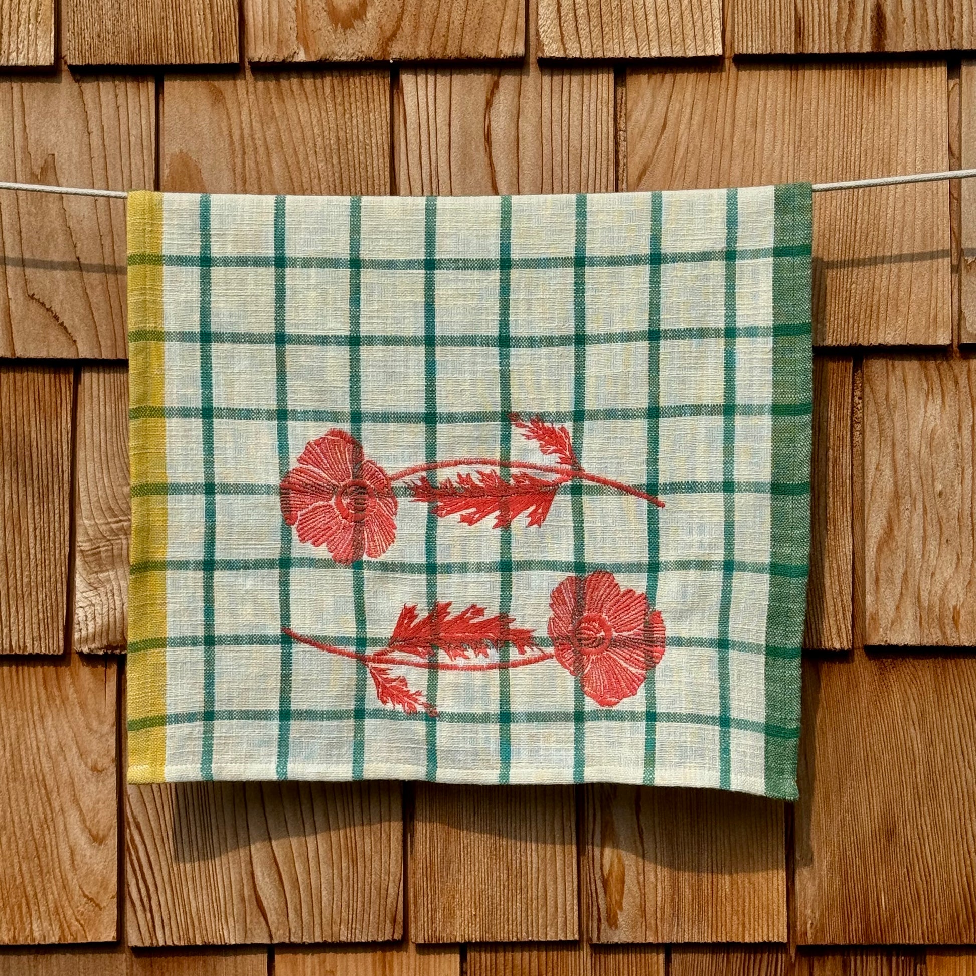 Cream and turquoise windowpane plaid tea towel with embroidered orange poppies, hanging on a clothesline