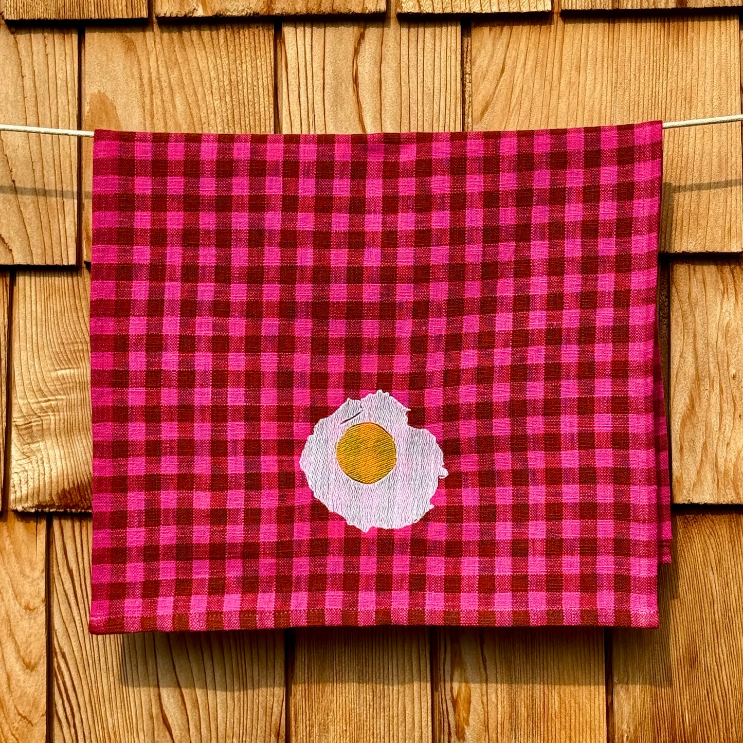 Fuchsia and cocoa gingham cotton tea towel with embroidered sunny-side-up egg, hanging on a clothesline