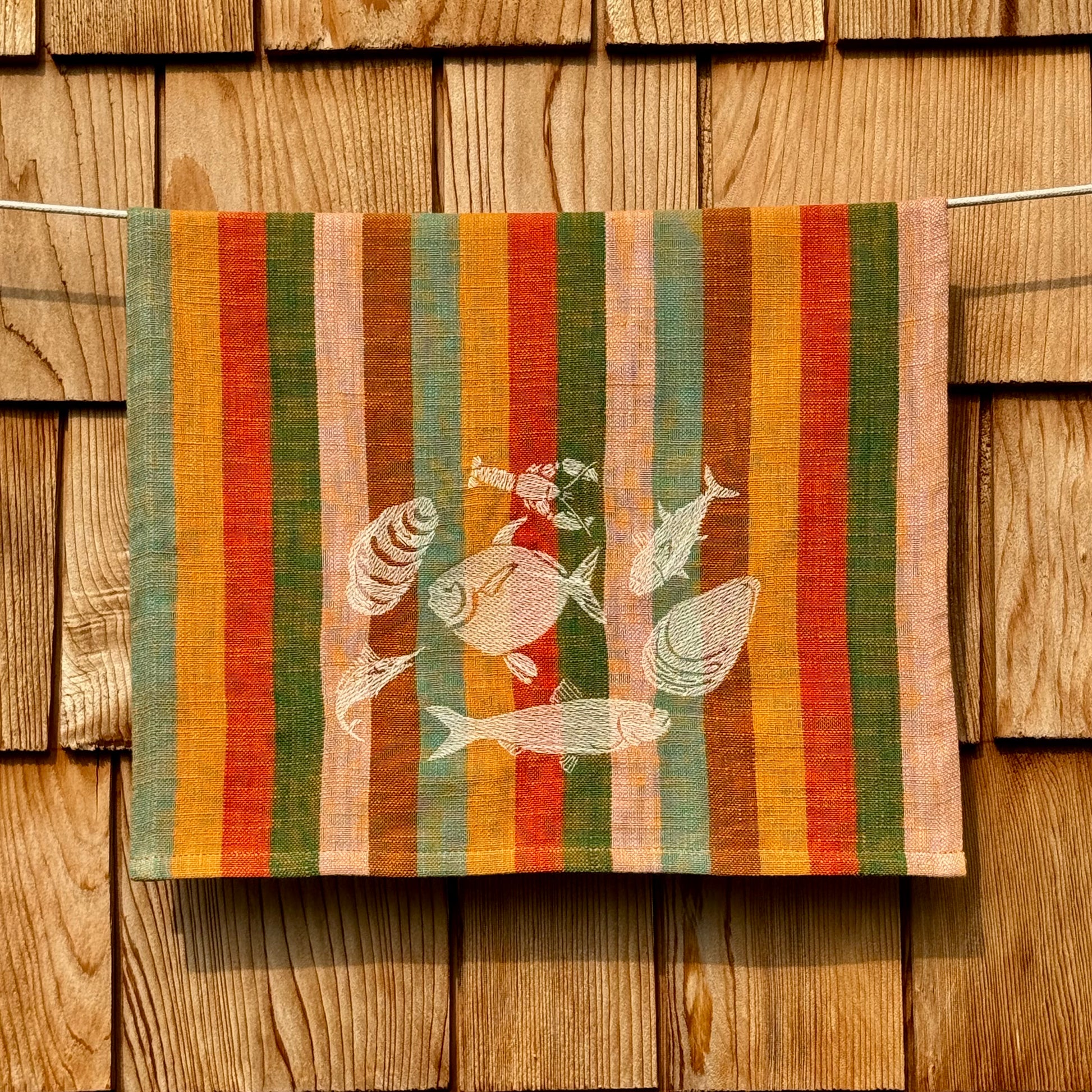 Multicolor striped cotton tea towel with cream embroidered fish and shell design, hanging on a clothesline