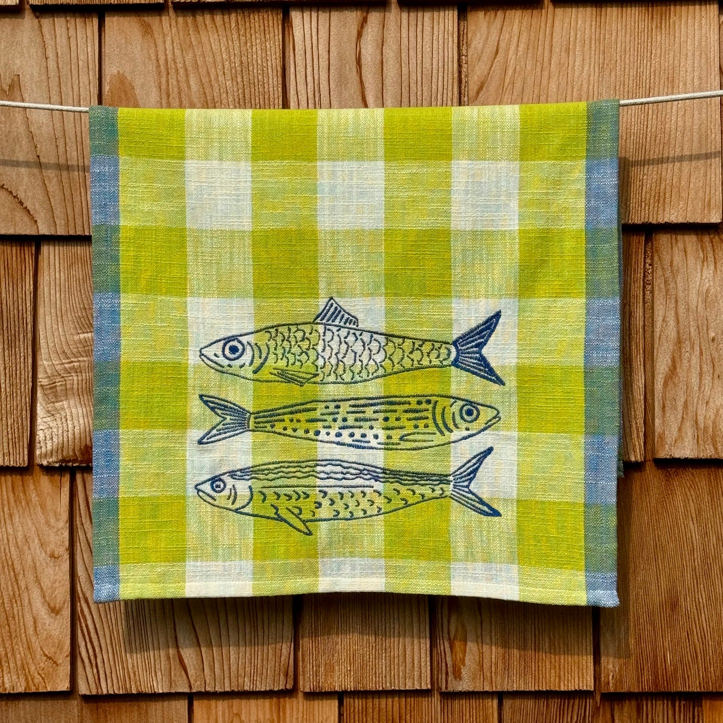 Citron, cream, and blue plaid cotton tea towel with three hand-embroidered fish, hanging on a clothesline