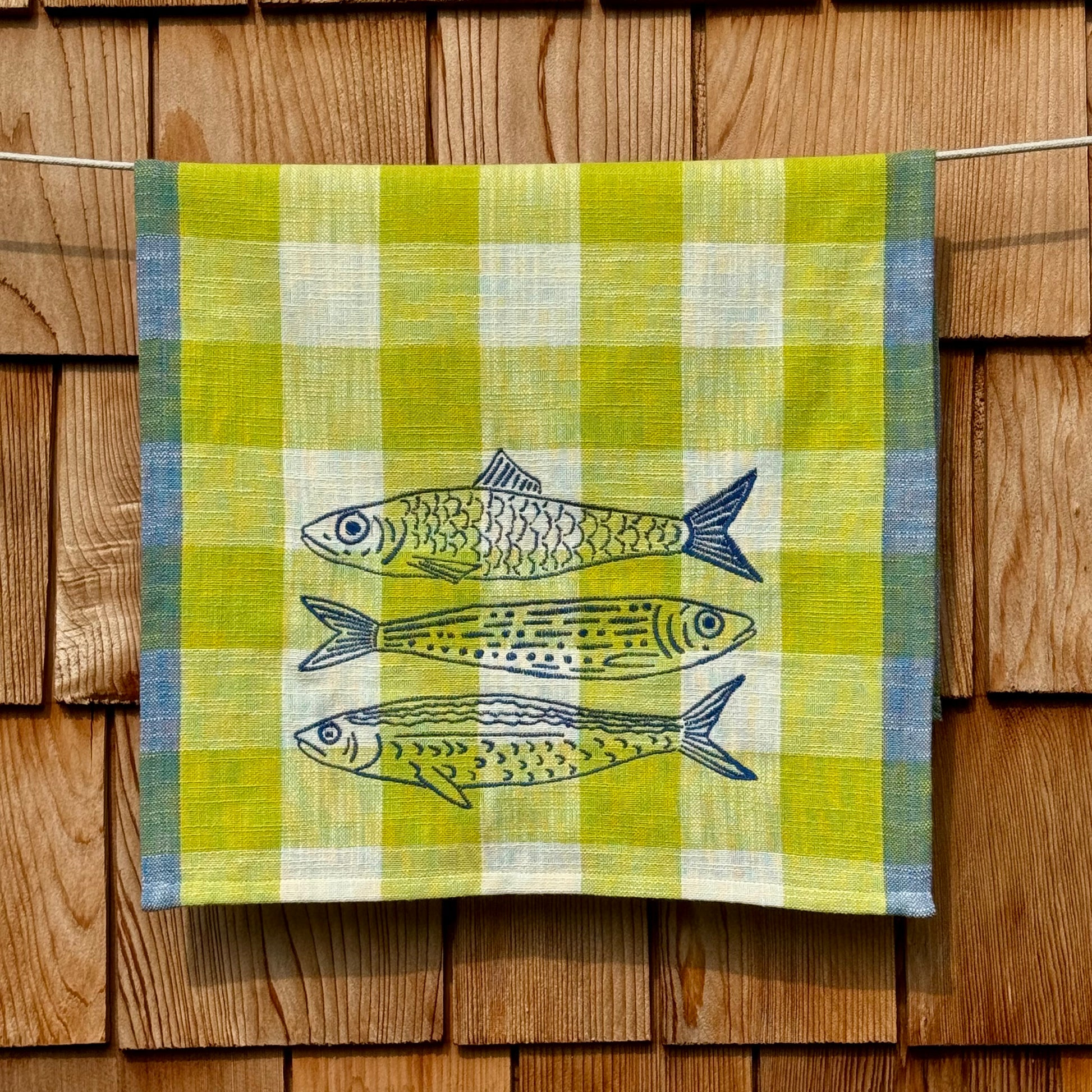 Citron, cream, and blue plaid cotton tea towel with three hand-embroidered fish, hanging on a clothesline